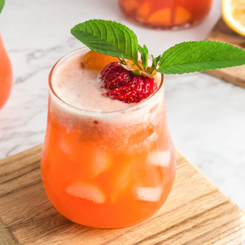Glass on wooden cutting board with white looking ice cubes and peach colored juice. It's topped with a sliced whole strawberry and 2 mint leaves.