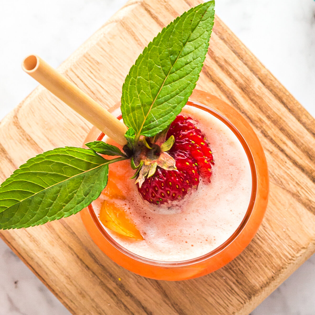 Looking down into the glass of frothy pink lemonade. There is a slice of peach, sliced strawberry, mint leaves and a gold straw sticking out. It's all on a small wooden board.