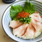 Sliced white hamachi sushi on rice with thinly sliced green onion, bright orange ikura fish roe and green shiso leaves in a light blue bowl.