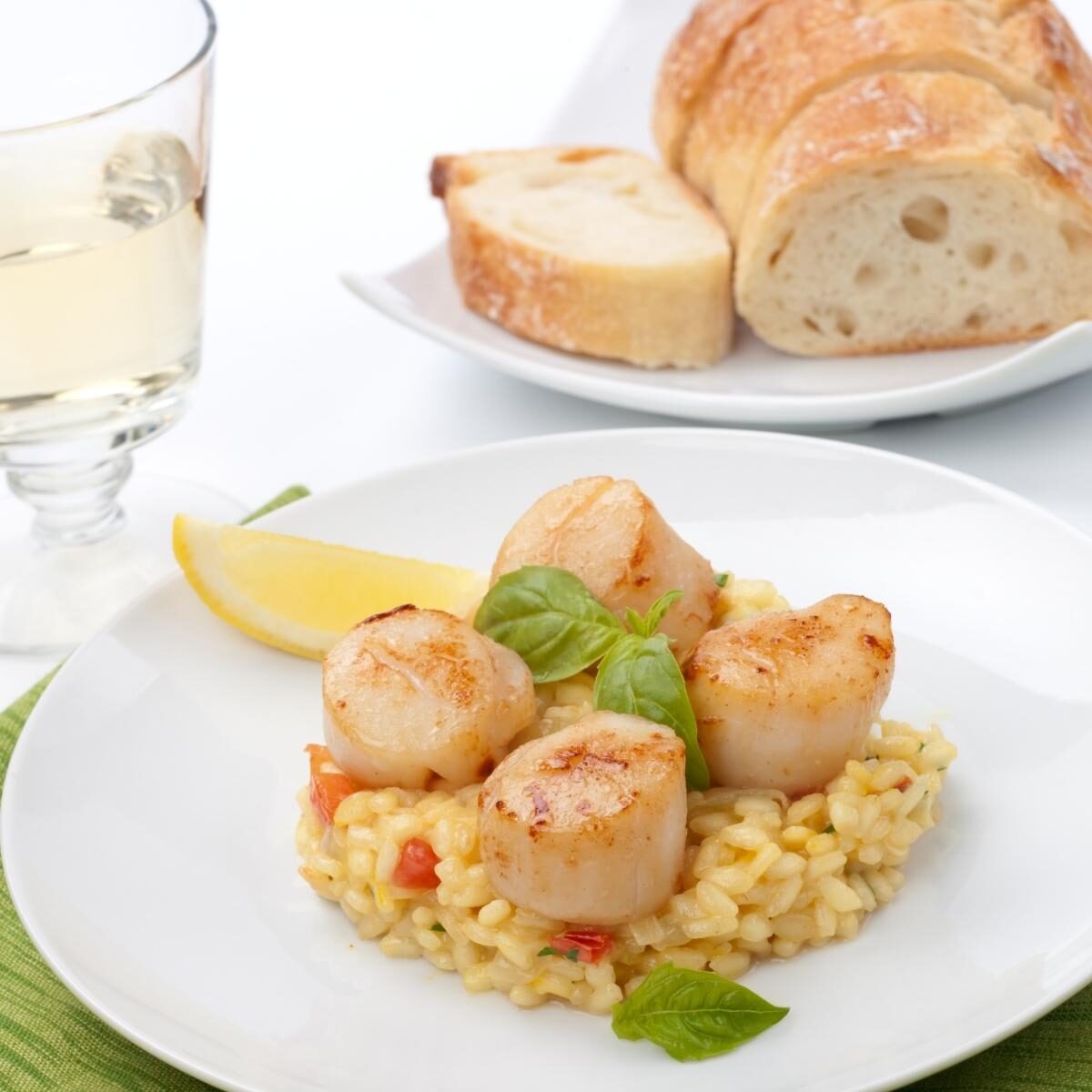 Seared scallops on creamy pale yellow risotto with a glass of white wine and sliced ciabatta bread in the background.