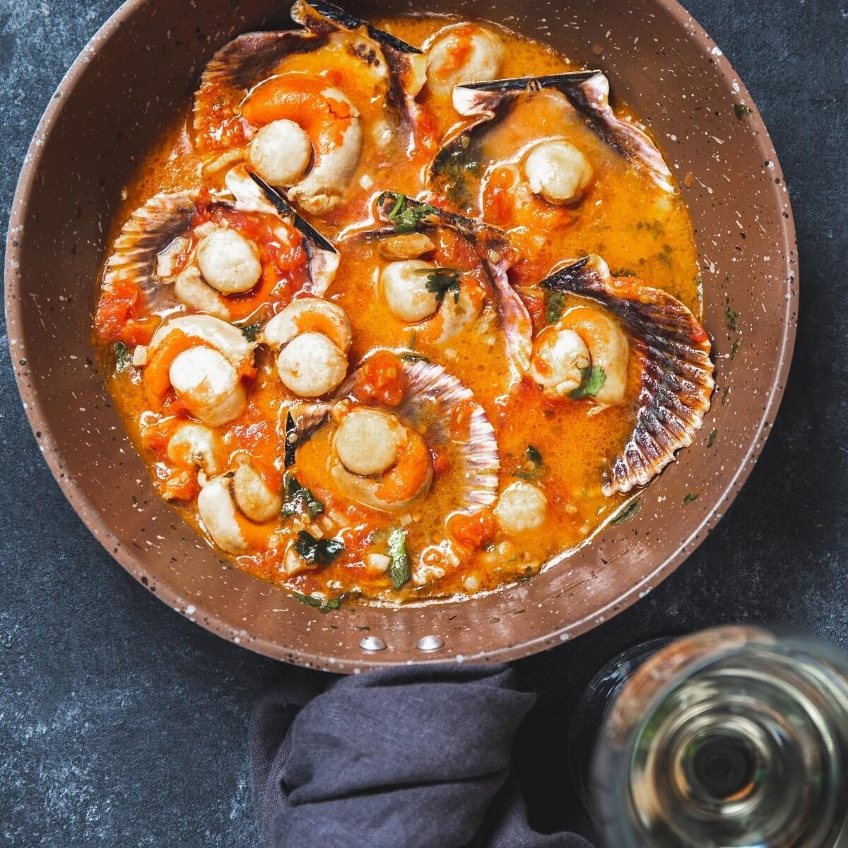 Deep red curry surrounding white round scallops sitting on their half shell in a brown bowl. There is a glass of white wine beside on the dark grey table.