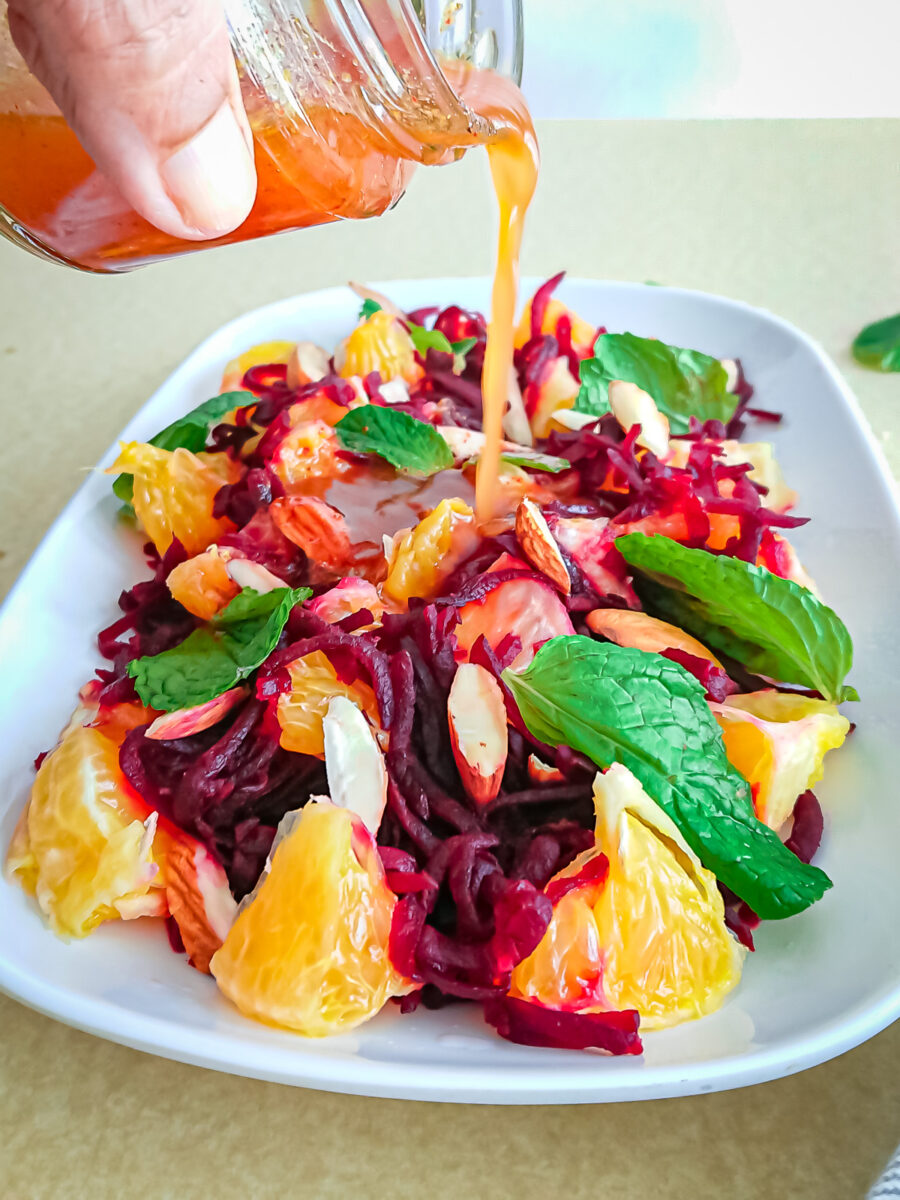 Bright red and orange beet and orange salad with green basil leaves scattered and an orange dressing being poured over from above.