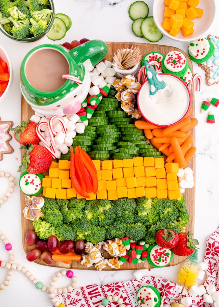 A wooden platter decorated to look like the green and brown buddy the elf sweater with broccoli, cheese cubes, carrots, marshmallows and grapes.