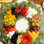 Edible wreath with simple cubes of cheddar cheese, balls of white mini bocconcini, green and red grapes, black and green olives and salami all on green leaves.