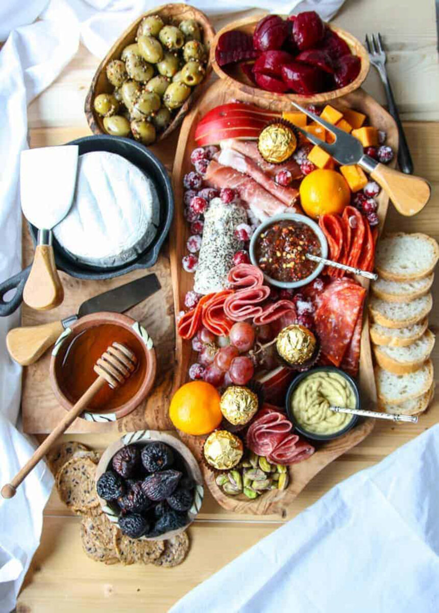 A wooden irregular shaped board with arranged cheese, meat, fruit, jam and olives.