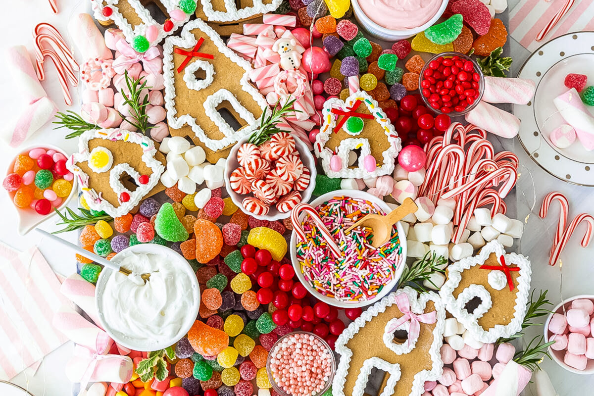 Board of candies and decorations with brown gingerbread house shaped cookies.