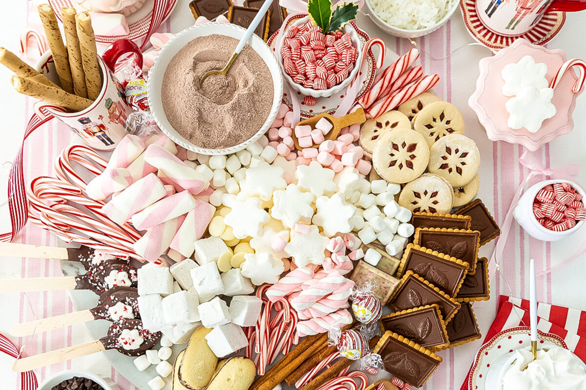 Stunning round board heaped with chocolate covered cookies, white and pink marshmallows, cany canes, hot chocolate powder mix, sugar cookies, whipped cream and cinnamon sticks.