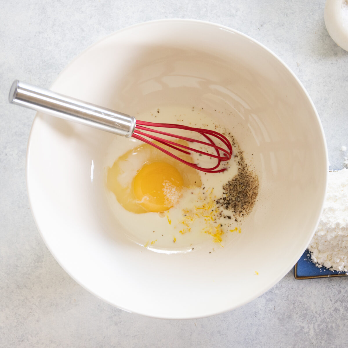 One egg, milk, lemon zest and pepper in white bowl with a silver and red whisk leaned against the inside of the bowl.