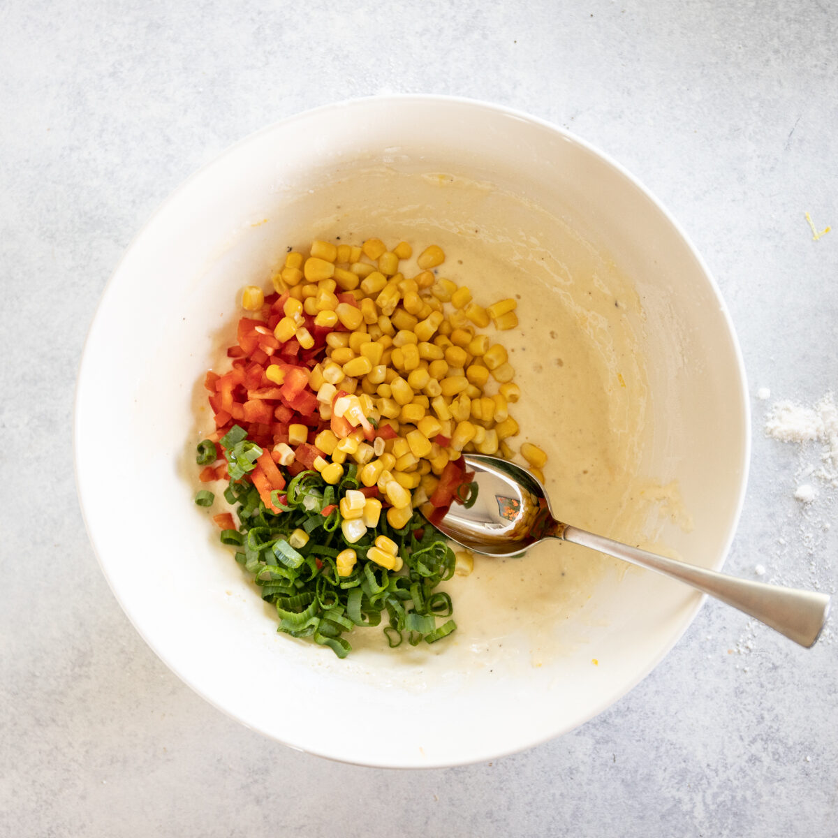 The cream colored batter has each ingredient poured in but not mixed yet. There is yellow corn kernels, diced red pepper and sliced scallions.