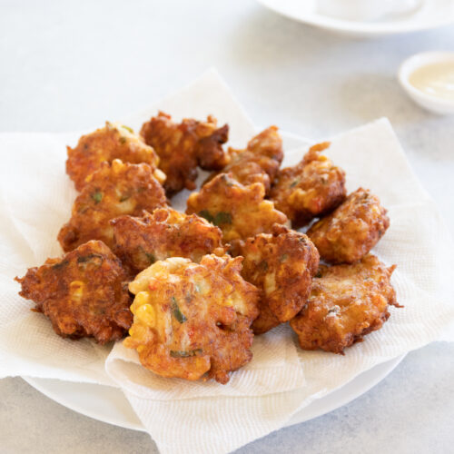 Paper towel lined plate full of crispy crab fritters on a white table with a small bowl of sauce in the background.
