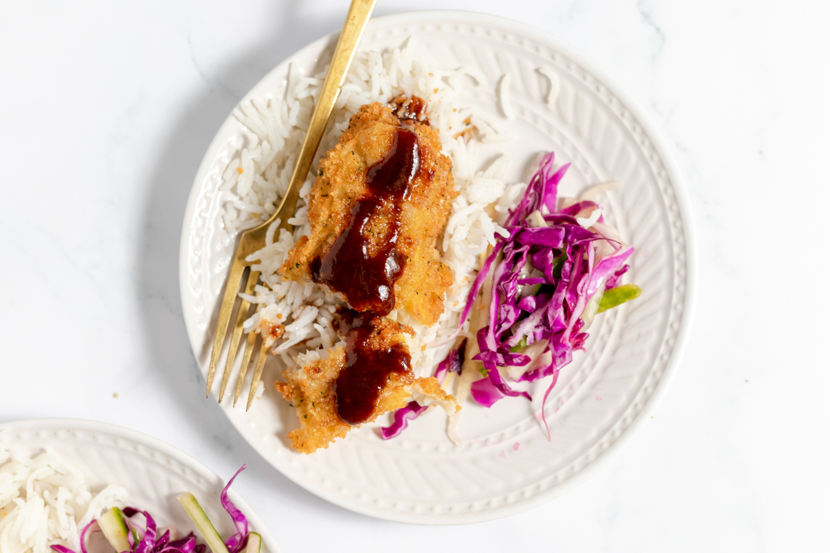 White plate on white table with golden crisp fried fish topped with a drizzle of dark brown sauce. There is shredded purple cabbage beside the fish on the plate.