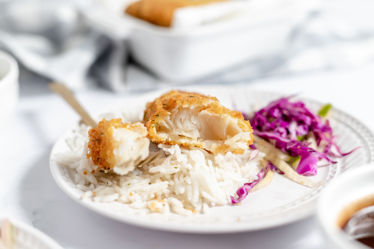White table and dishes with a plate of white rice, shredded red cabbage and crispy fish that has been broken open to expose the moist and flaky white interior.