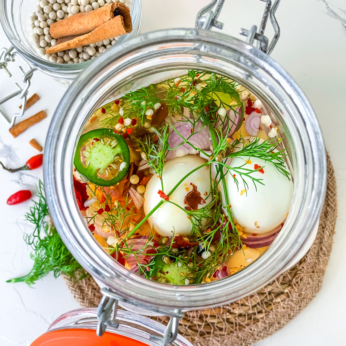 Overhead view of peeled hard eggs in mason jar with mustard seed, sliced green jalapenos, herb fronds and red pepper flakes all in vinegar mixture. 