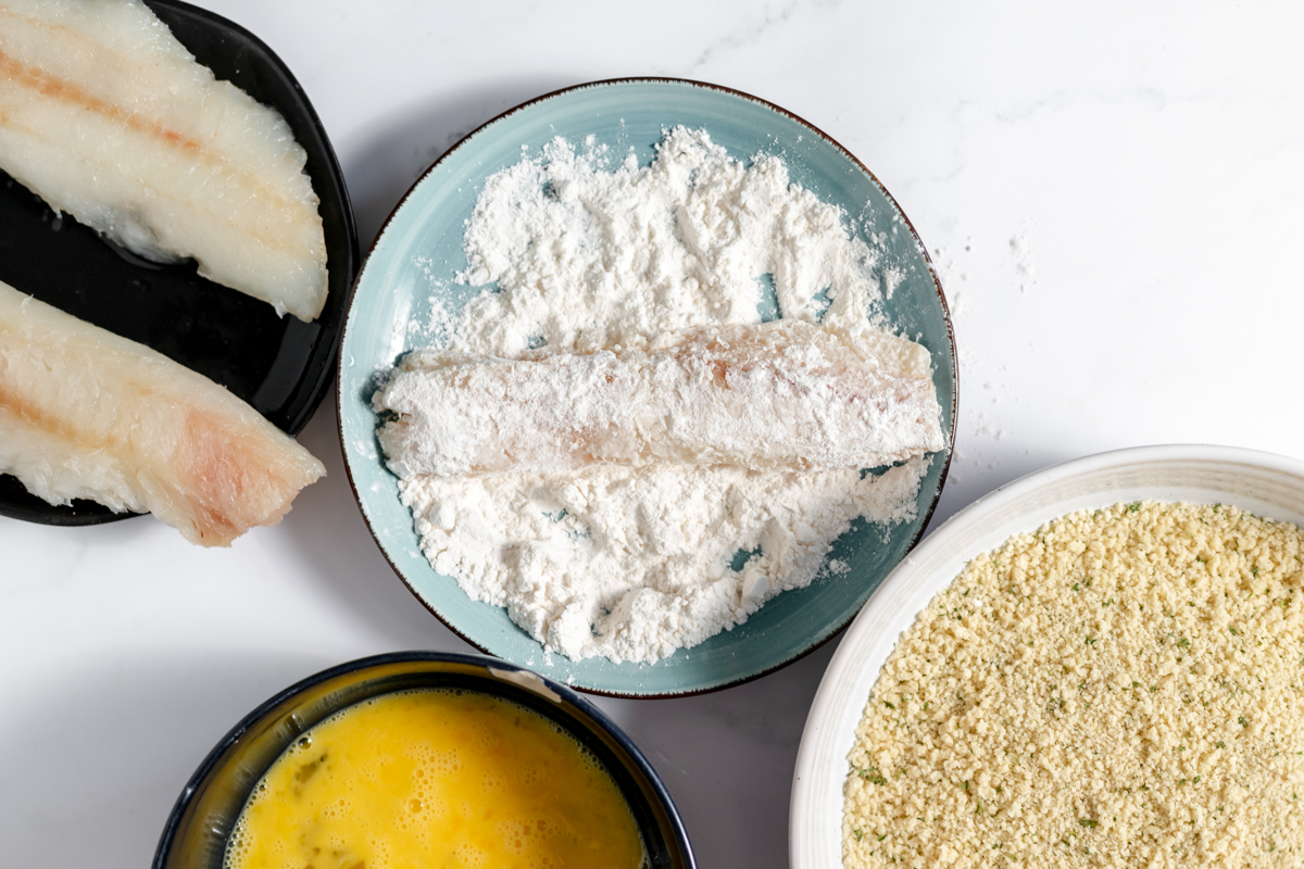 One fish filet in coated in flour on a blue plate beside the yellow egg wash, cod filets and panko bread crumbs.