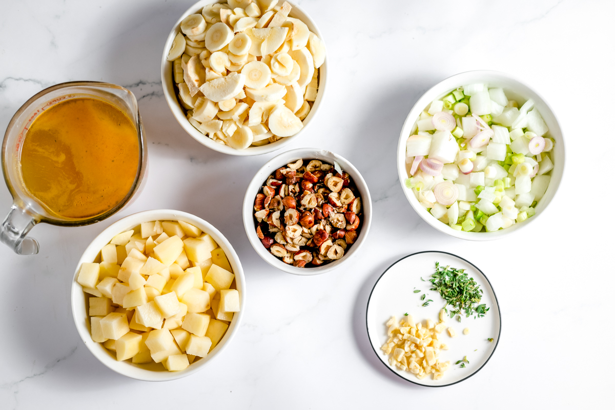 Bowls of each of the prepared ingredients. There is sliced rounds of parsnip, diced potato, chicken stock, hazelnuts, garlic, thyme and pale green onions.