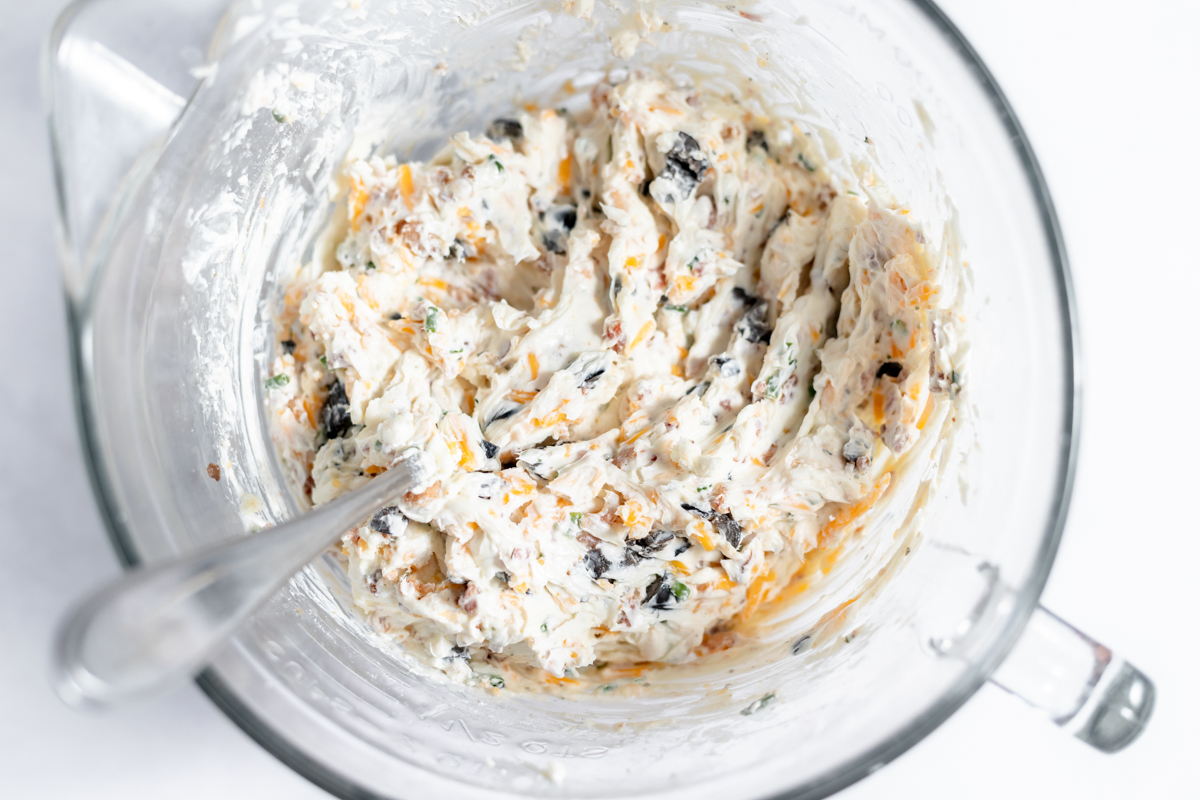 Cream cheese mixture has been mixed in the glass measuring cup bowl. It is white with flecks of orange, green and black.