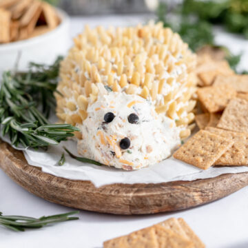Front view of the hedgehog with crackers and rosemary.d mouth. It is sitting on a parchment covered cutting board and surrounded with green rosemary sprigs and square crackers.