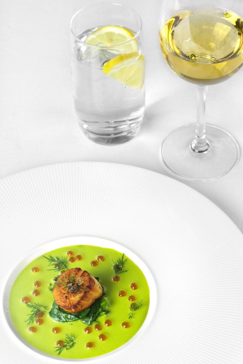 White plate with green sauce and golden seared scallop in the middle with water and white wine in the background. 