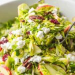 Bright green shaved brussels sprouts with brown nuts, red and white sliced apples and white crumbled feta cheese in a white bowl.