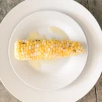 White plate with corn on the cob on it and melted butter on and around the yellow corn.