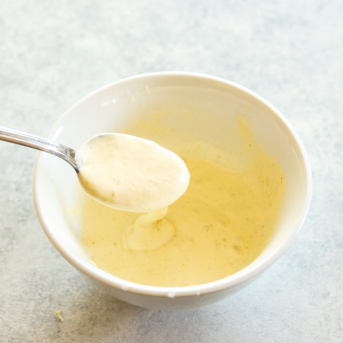 White bowl of creamy pale yellow sauce. There is a spoon lifting some out and it is dripping back into the bowl.