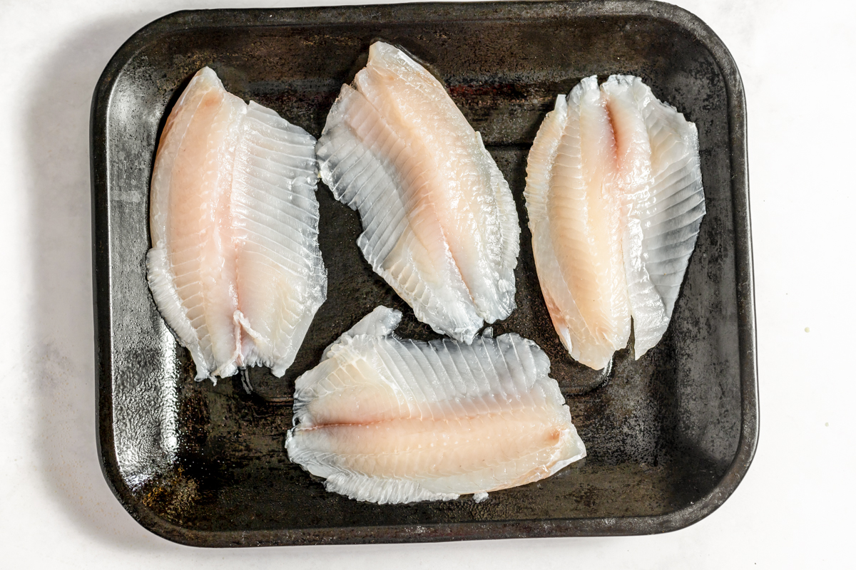 A black baking tray with four white tilapia fillets spread on it.