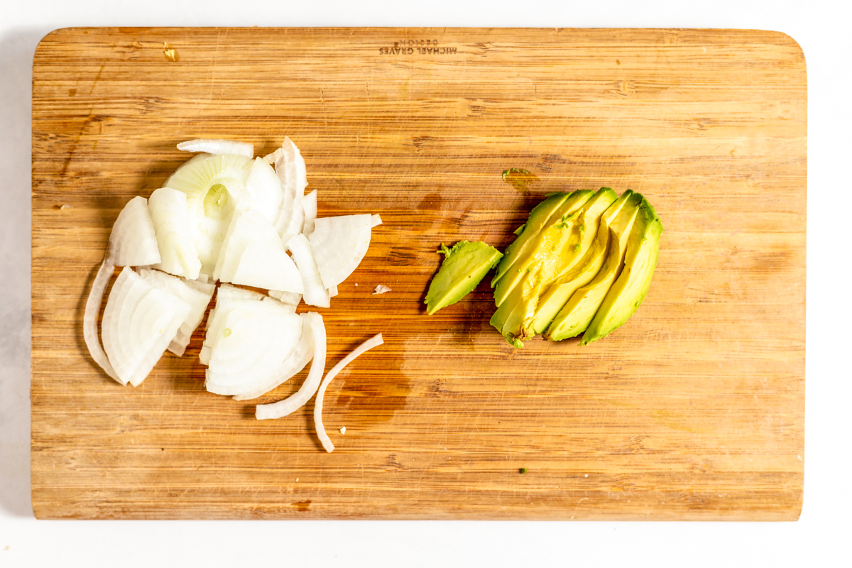 A wooden cutting board with sliced white onion and avocado on it.