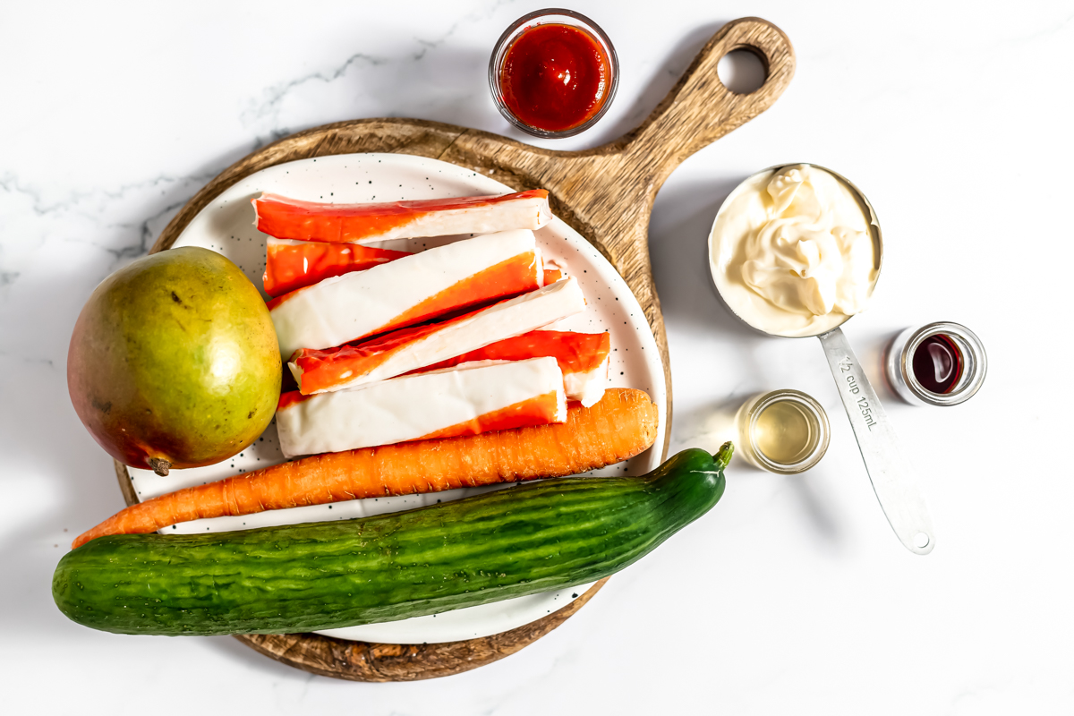 Ingredients including kani sticks, cucumber, carrot, Mango, hot sauce, and mayo on a cutting board on a white table.