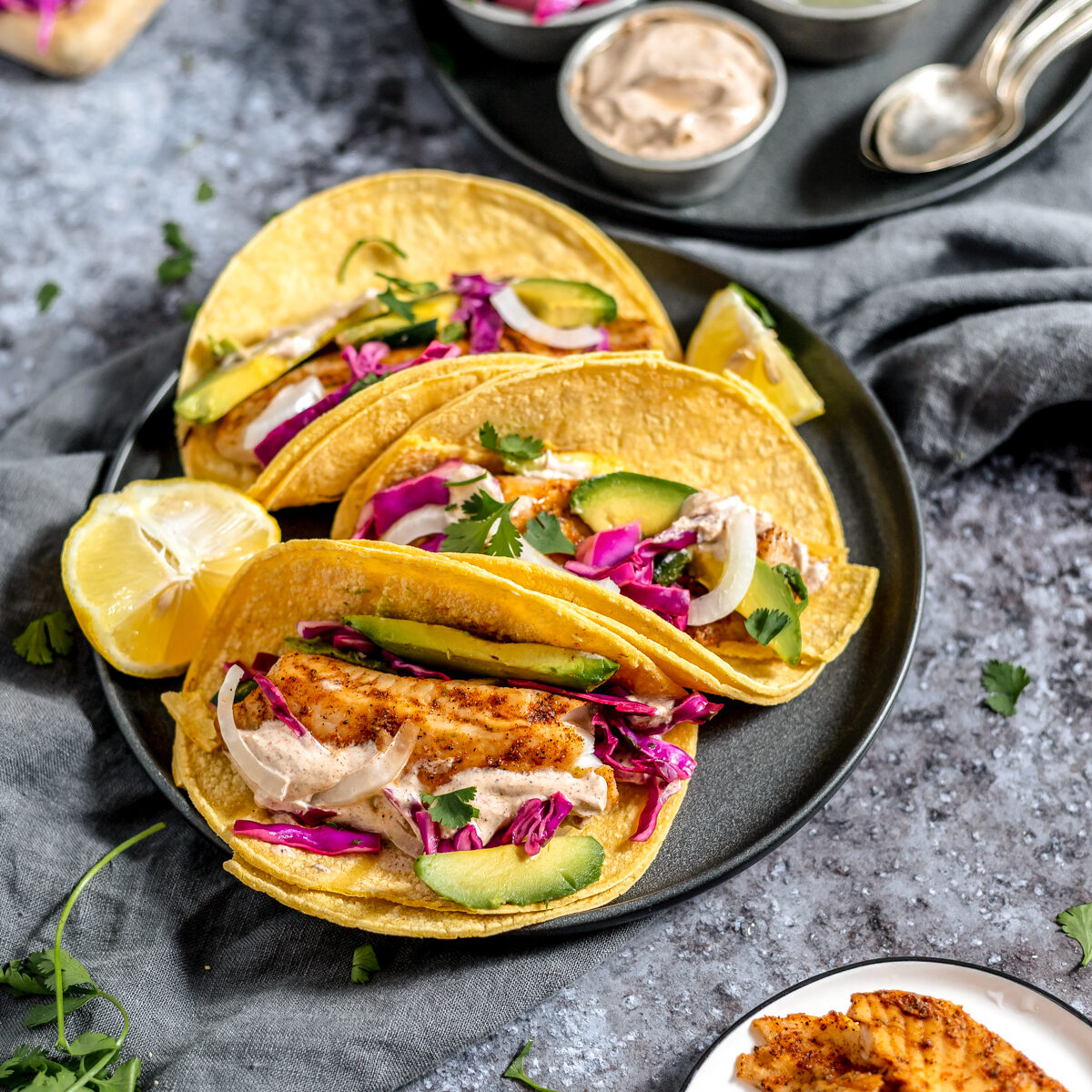 Three yellow corn tortillas folded in half and stuffed with orange fish, green avocado, purple cabbage and creamy sauce on a grey plate set on a grey countertop.