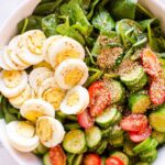 Green spinach salad with sliced eggs arranged on one third and tomatoes and cucumber arranged on another third. It is in a white bowl on a white table.