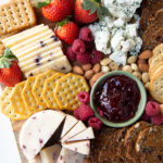 Square wooden board laden with strawberries, assorted cheese and crackers, jam and nuts.