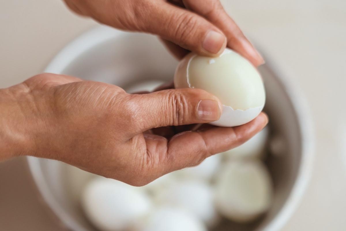 Hands peeling a white egg over a silver pot of eggs and water.