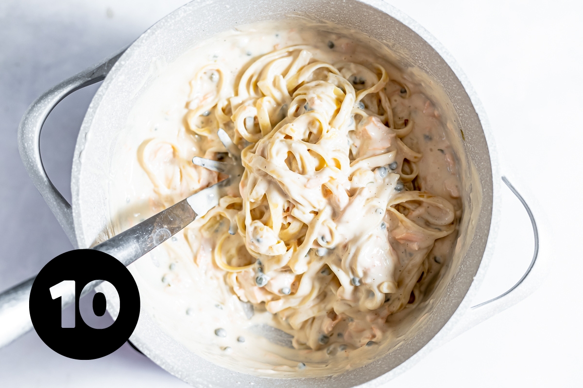 Fettuccine is getting stirred into the thick creamy sauce with a large metal spoon.