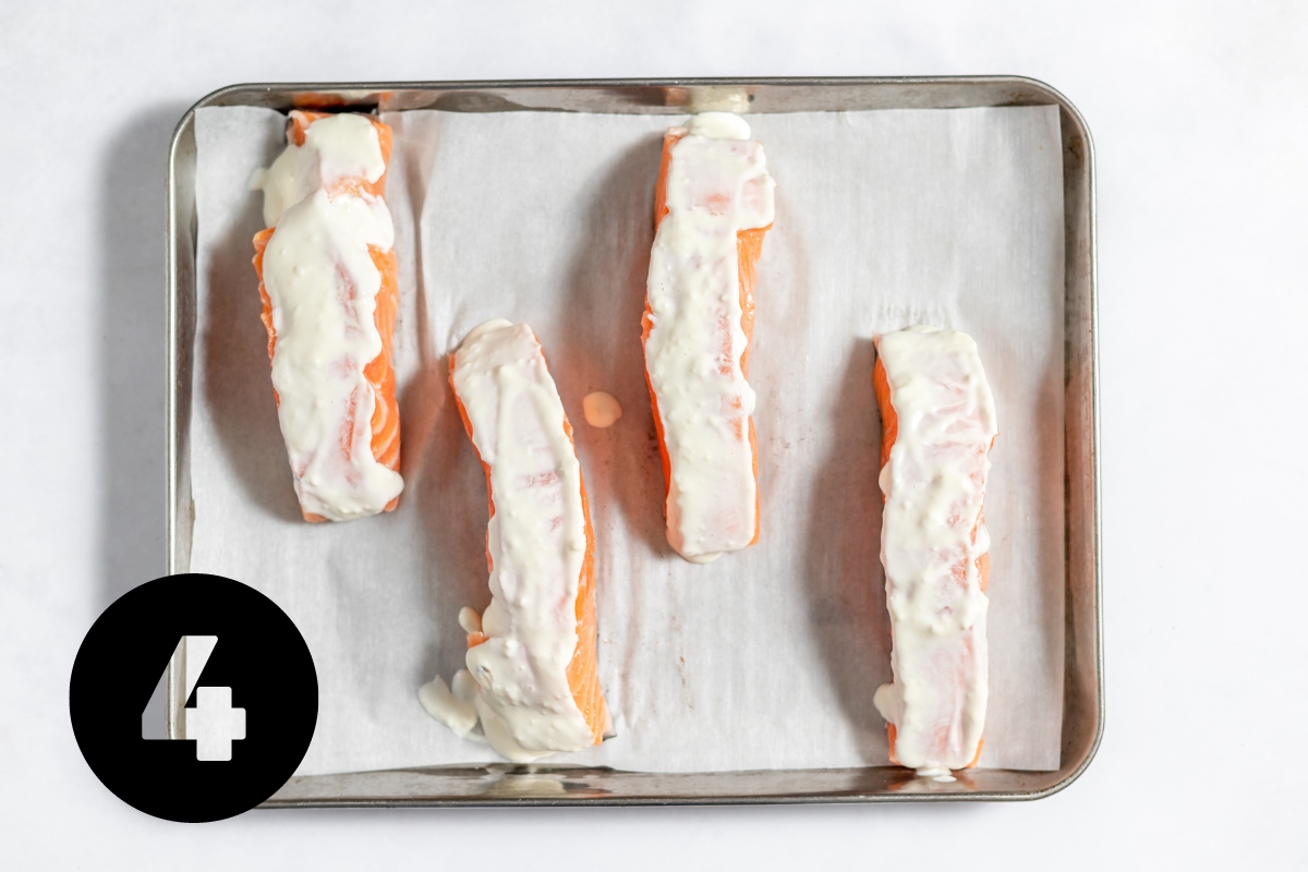 Parchment paper lined baking tray with 4 rectangular pieces of salmon covered in white mayonnaise mixture.