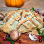 A braided pastry Wellington on a cutting board surrounded by mushrooms and cranberries.