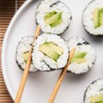 White and green slices of an avocado sushi roll. One piece is held up close with wooden chopsticks.