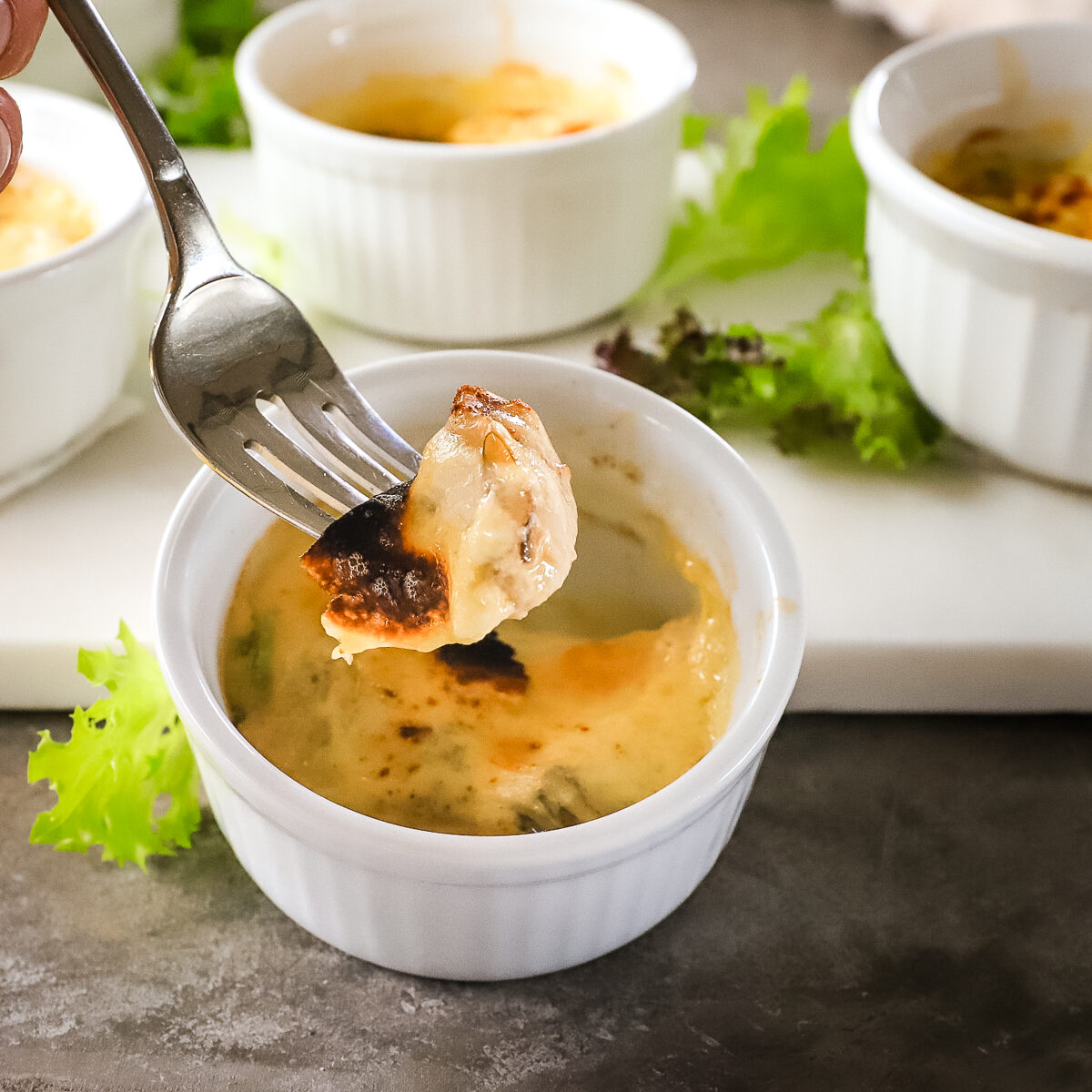 A fork with a piece of cheesy baked oyster lifted out of a white ramekin. It is a creamy yellow color with bubbly black and brown spots on top. 