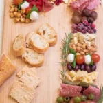 Candy cane shaped charcuterie on a wooden board. Sliced meat, grapes, tomatoes and bocconcini make up the cane and bread slices are served beside.