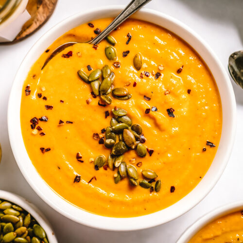 White bowl full of bright orange smooth soup topped with pepitas. A spoon in the bowl and more spoons beside.