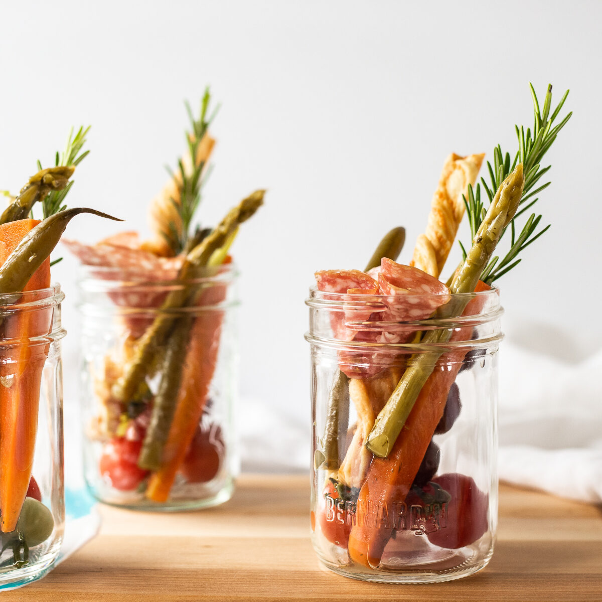 3 Small mason jars filled with sliced meat, pickled vegetables, bread sticks and rosemary sprigs.