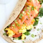 Whole wheat tortilla partially wrapped around scrambled eggs and thinly sliced smoked salmon with chives and a smear of cream cheese inside.