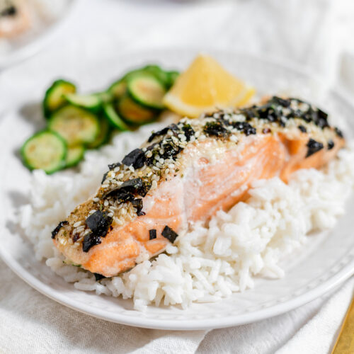 White round plate of white rice topped with pink colored salmon topped with seaweed and sesame seed mixture with a side of cucumber salad.