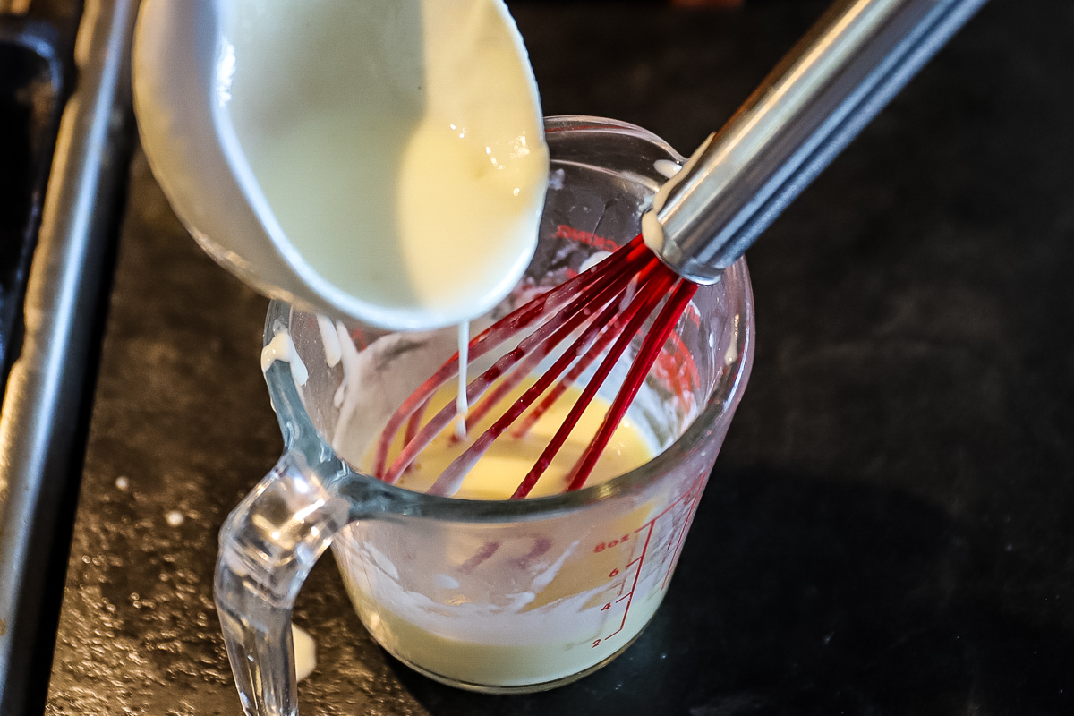 Hot white bechamel sauce being slowly poured into the measuring cup with the egg yolk in it.