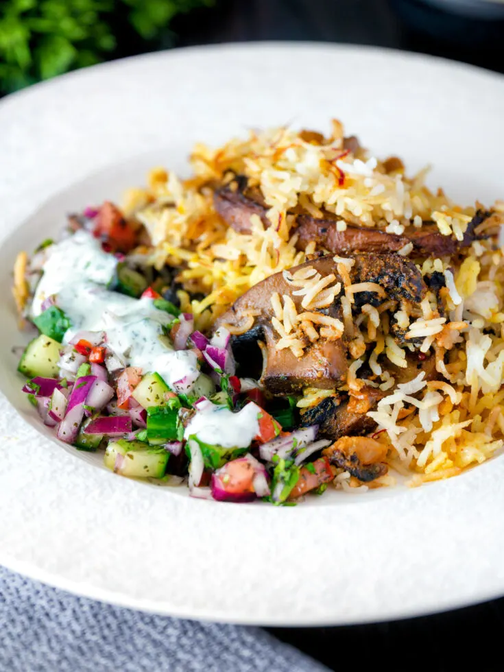 Bowl of biryani topped with mushrooms beside green and red salad topped with yogurt.