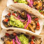 Three soft tacos side by side with brown mushrooms, purple pickled onions and green avocado in them.