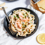 Black bowl of white and pink fettuccine noodles with cream sauce and flaked salmon in a black bowl on a white tablecloth. Overhead view with lemon slices to the bottom right.