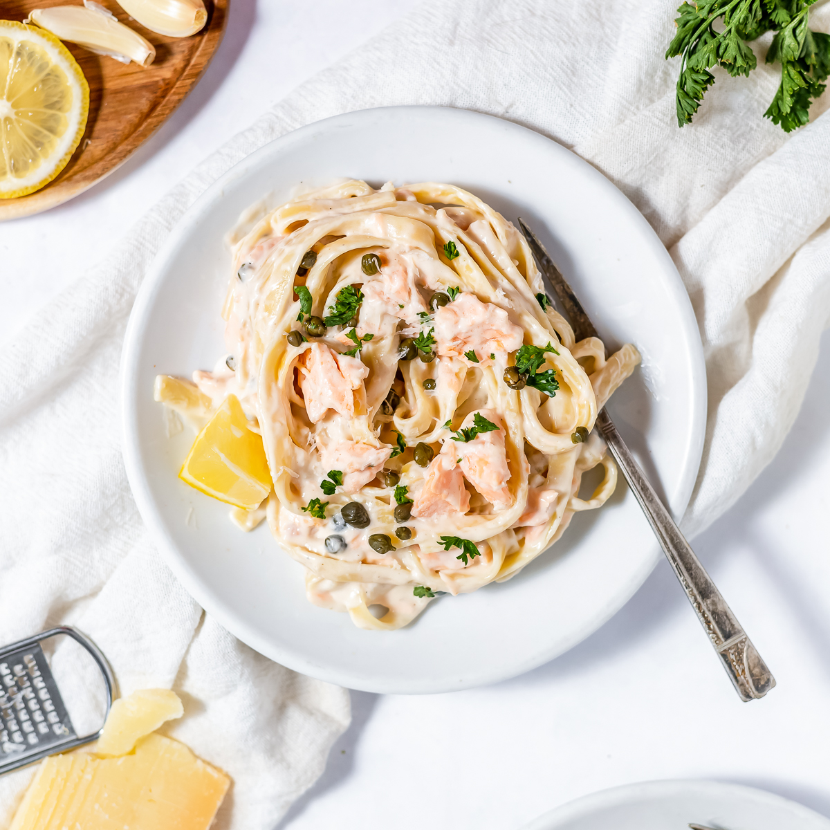 White round bowl of creamy fettuccine with pink pieces of salmon, dark green capers, chopped parsley sprinkled over and a small wedge of lemon beside.