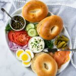 A platter of bagels, smoked salmon, capers, cream cheese, lemon wedges, red onion, sliced hard boiled egg, and cucumber.
