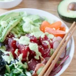 White bowl filled with sushi ingredients including cubes tuna, chopped green onion, radish and avocado with a pale green creamy sauce drizzled over.