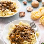 Bowls of flat wavy noodles topped with mushroom stroganoff mixture on top. Mushrooms and fresh pasta are sprinkled around the table between the bowls.